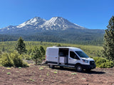 A white Ford Transit RV rental van, door open, sits on dirt, facing snow-capped Mount Shasta & forested valley under blue sky.