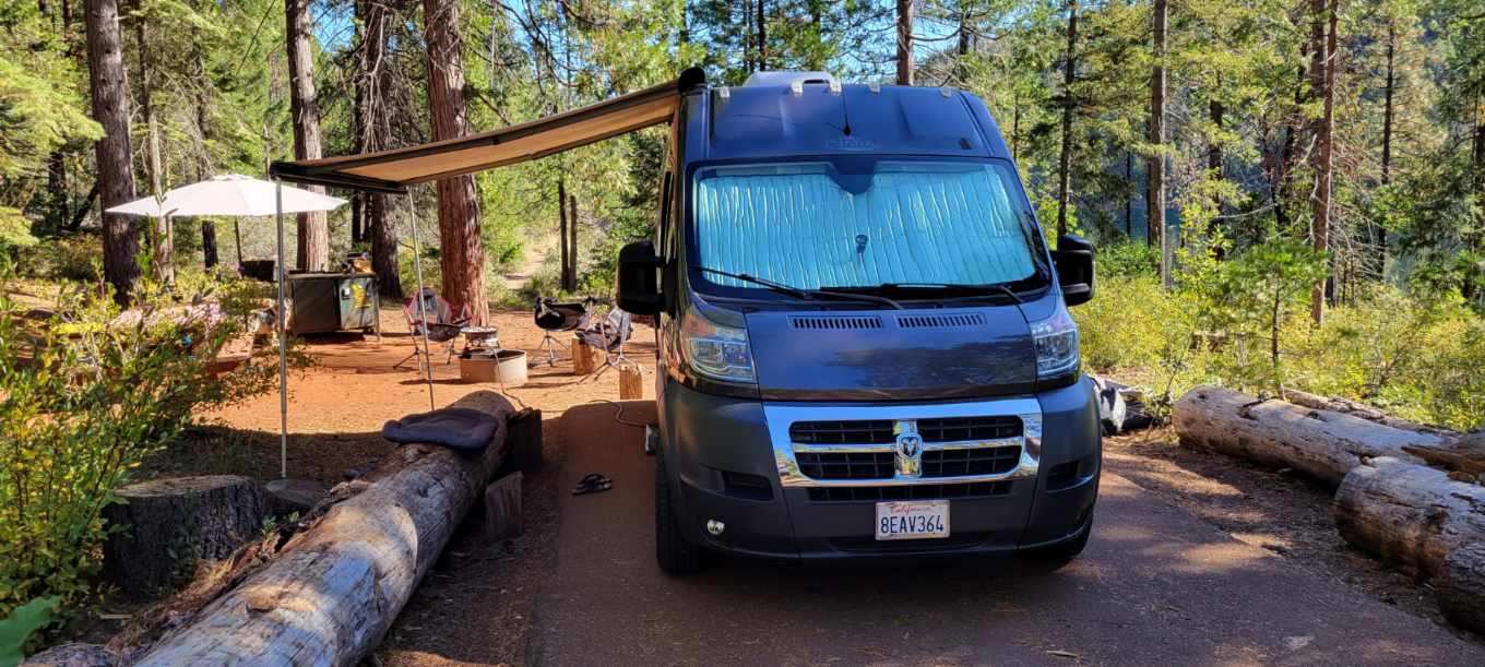 This RV rental camper van is set up at a forest campsite with an awning, sunshade, and comfortable outdoor living space. — photo 16