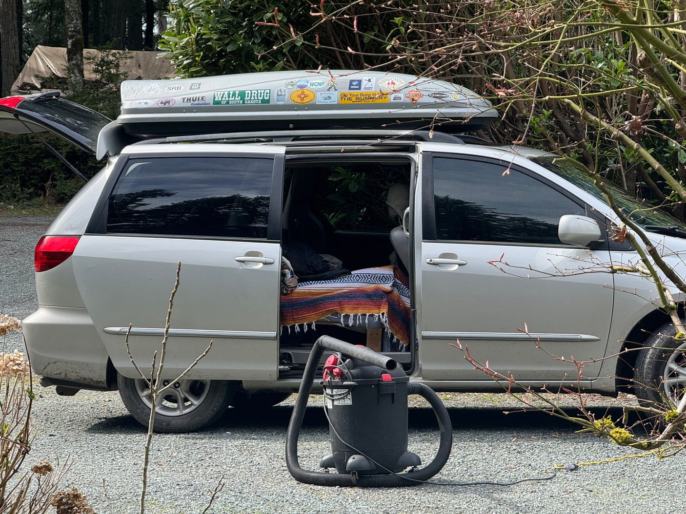 Minivan camper, like an RV rental, with an open side door showing a bed, a roof cargo box, and a shop vac for outdoor cleaning.