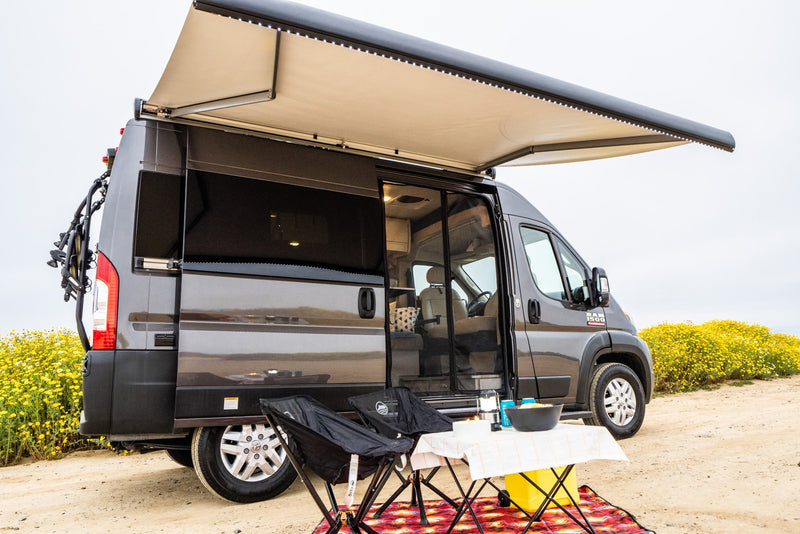 RV rental camper van with open side, extended awning, and outdoor camp setup on a sandy path next to yellow flowers. — photo 4