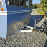 Light blue RV rental trailer, featuring white stripes, hitched to a car on a sunny gravel driveway.