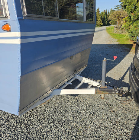 Light blue RV rental trailer, featuring white stripes, hitched to a car on a sunny gravel driveway.