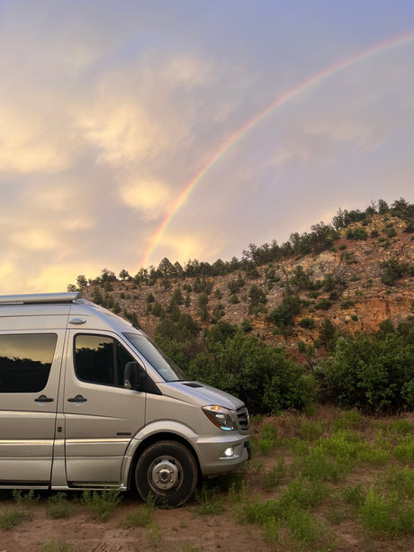 A silver rv rental van parked by a rocky, tree-lined hill under a vibrant rainbow and warm, cloudy sky.