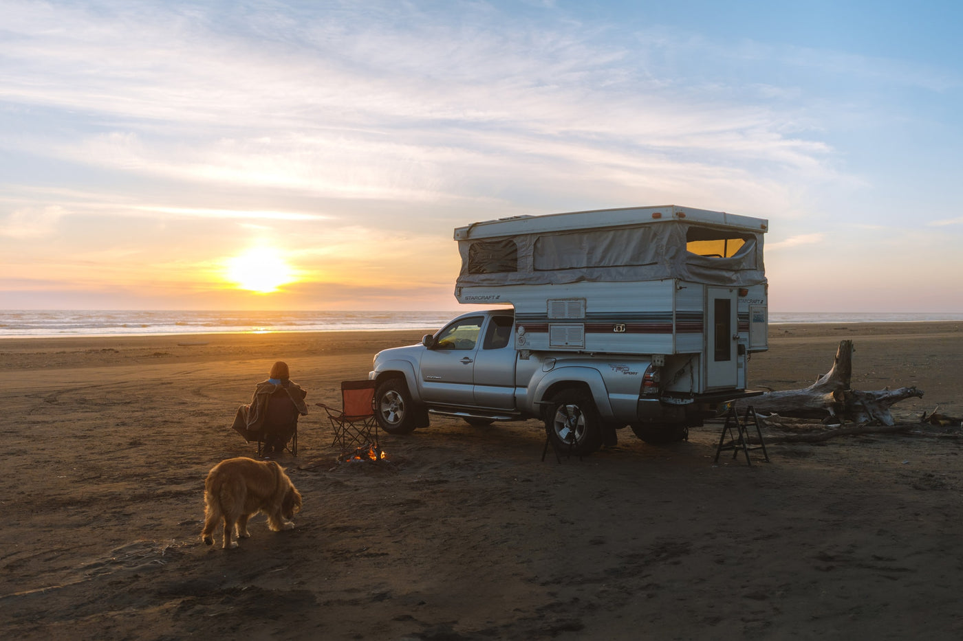 Truck camper RV rental parked on a sandy beach at sunset. A person and dog relax by a crackling campfire near the ocean. — photo 2