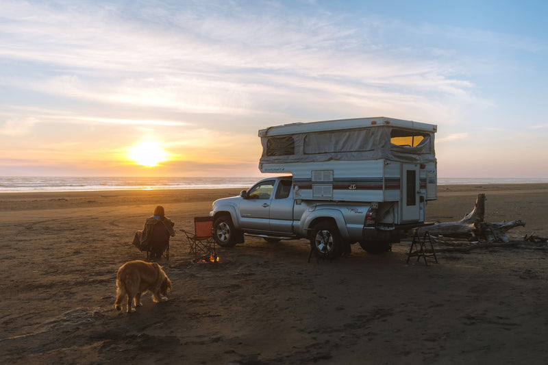 Truck camper RV rental parked on a sandy beach at sunset. A person and dog relax by a crackling campfire near the ocean. — photo 2
