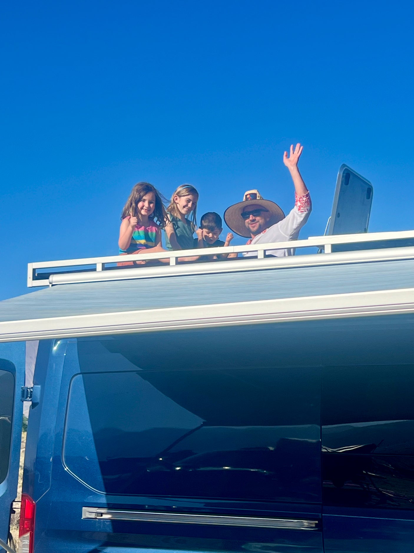 Family enjoying their RV rental: an adult and three children wave and smile from the blue RV's rooftop against a clear sky.