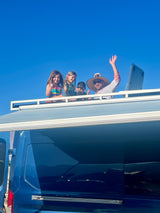 Family enjoying their RV rental: an adult and three children wave and smile from the blue RV's rooftop against a clear sky.