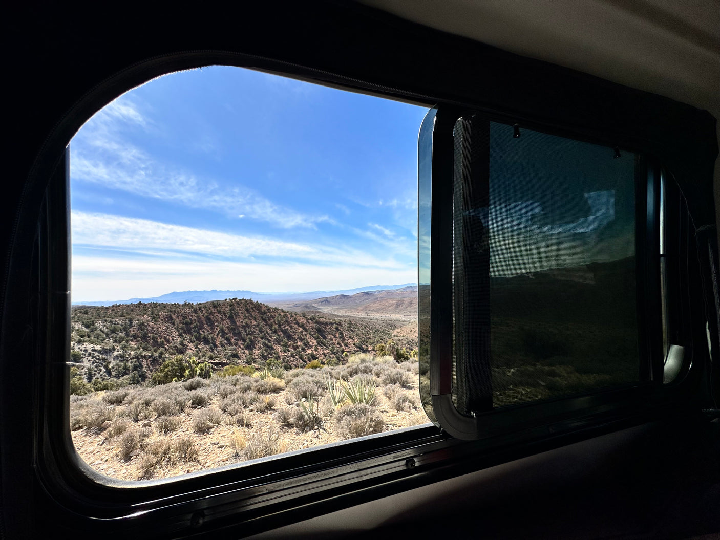 Framed by an RV rental window, a bright desert landscape with mountains and blue sky stretches. Inside, a dark interior and shaded screen.