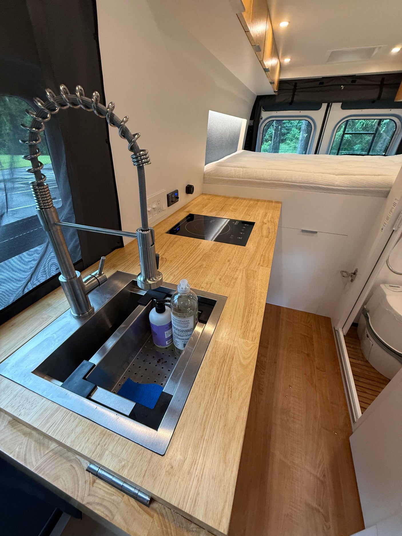 Inside a sleek rv rental: a compact kitchen with stainless steel sink & induction cooktop, leading to a cozy elevated bed.