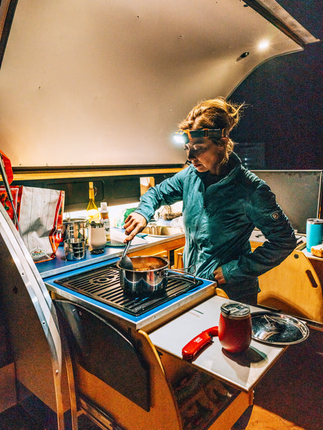 Inside an RV rental, a woman with a headlamp cooks dinner on a portable stove in her compact kitchen.
