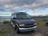 A blue Ford van, perfect for an RV rental adventure, parked on an Alaskan rocky beach facing snow-capped mountains.