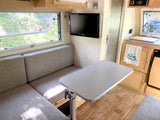 Inside a compact rv rental: dinette, small kitchen, and TV. Bright window view of nature.