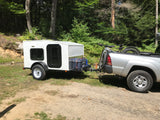 A white teardrop trailer (like an RV rental) is hitched to a silver pickup truck with mountain bikes, parked in a wooded area.