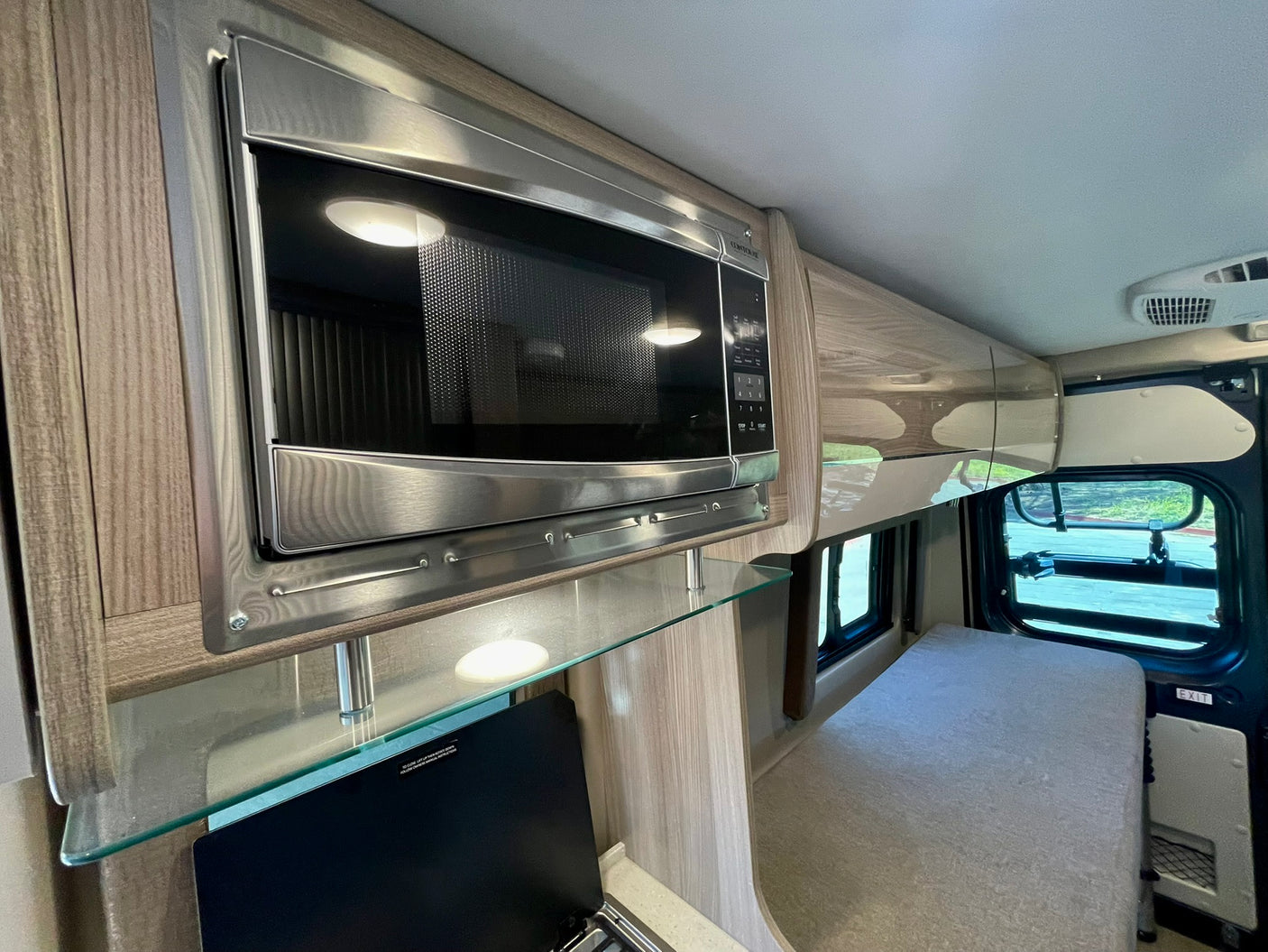 Modern RV rental interior with stainless steel microwave, glass shelf, and compact bed area near a window.