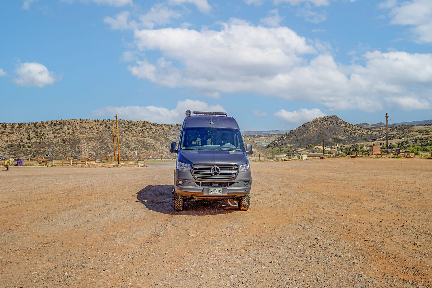 A gray Sprinter RV rental parked in a vast, dusty outdoor lot with arid mountains and a blue, cloudy sky in the background.