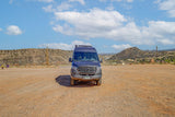 A gray Sprinter RV rental parked in a vast, dusty outdoor lot with arid mountains and a blue, cloudy sky in the background.