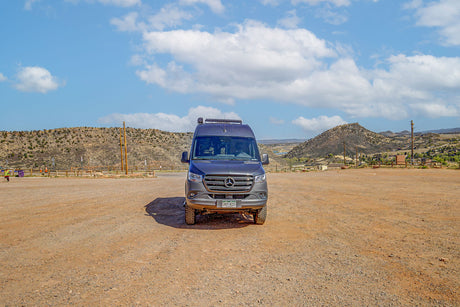 A gray Sprinter RV rental parked in a vast, dusty outdoor lot with arid mountains and a blue, cloudy sky in the background.
