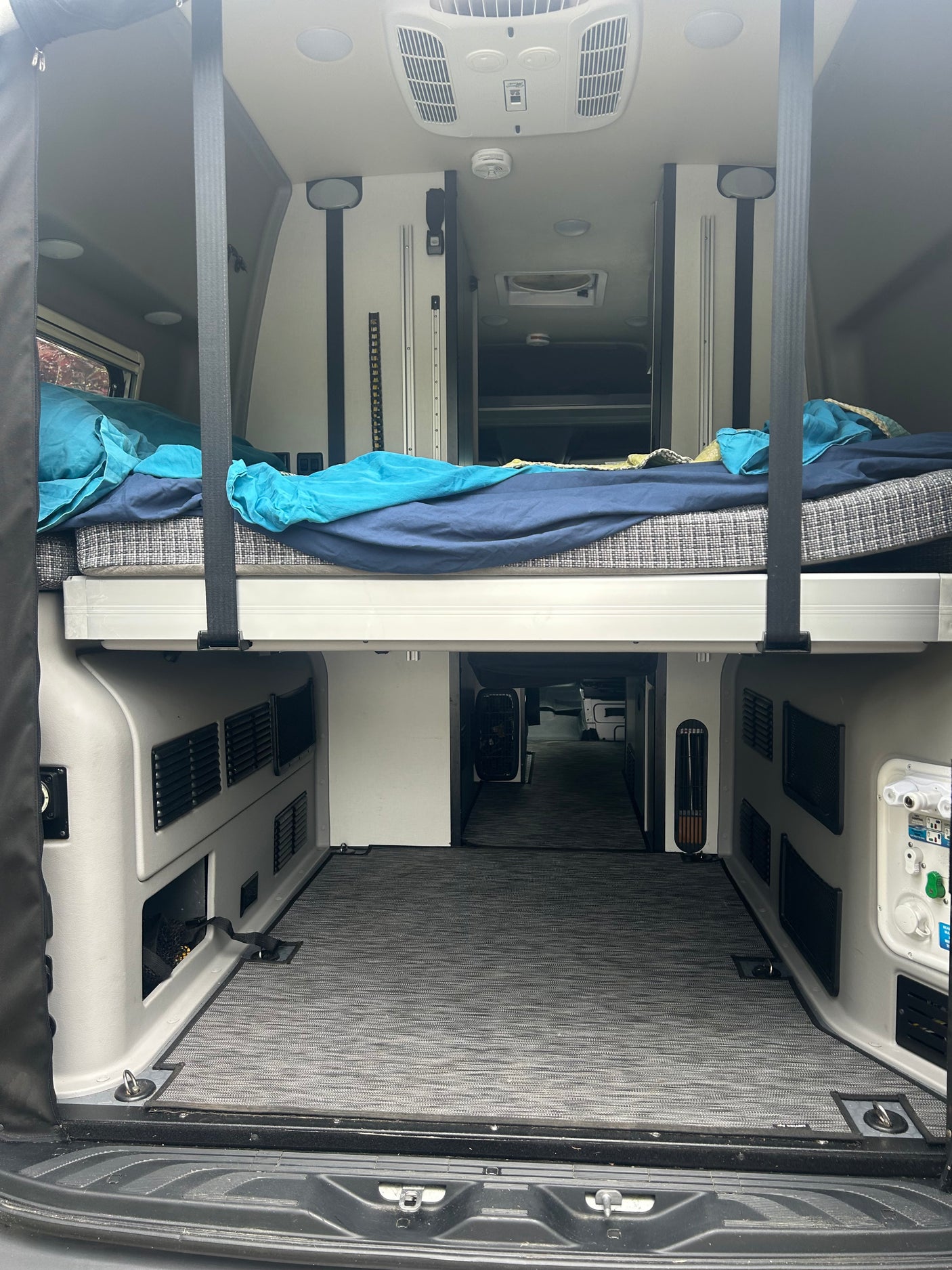 Rear interior of an rv rental camper van showcasing a raised bed with bedding, ample under-bed storage, and a well-equipped ceiling.