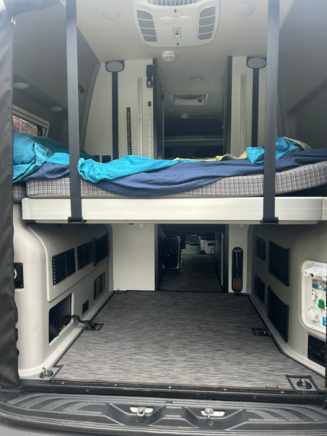Rear interior of an rv rental camper van showcasing a raised bed with bedding, ample under-bed storage, and a well-equipped ceiling.