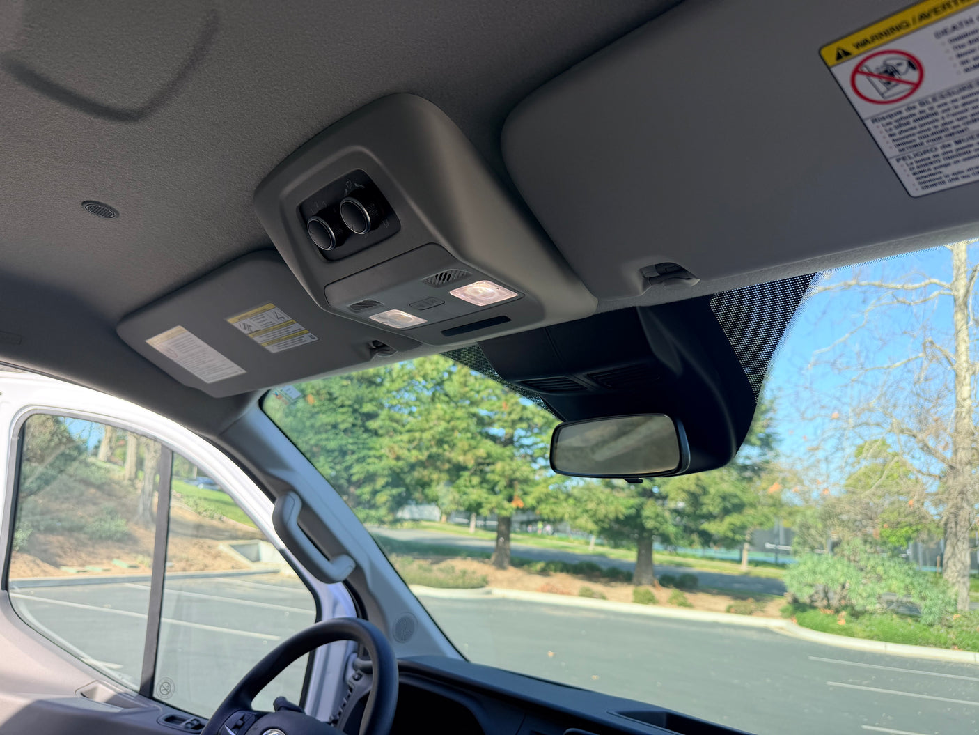 RV rental van cabin: Overhead console with controls & lights, sun visors, and rearview mirror inside, with trees outside.