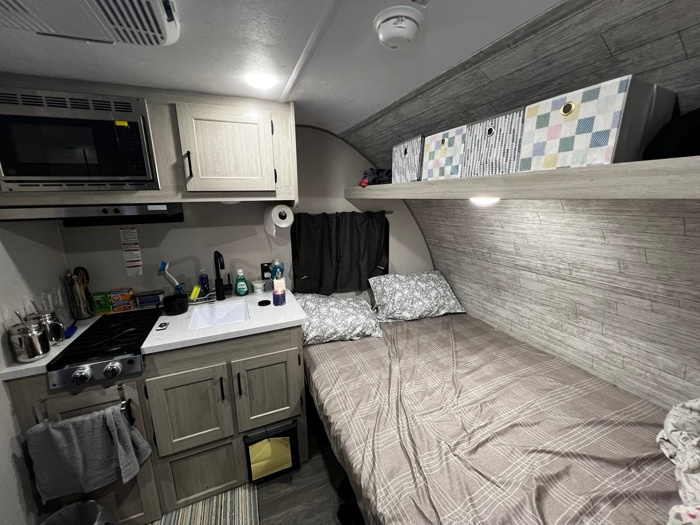 RV rental interior: compact kitchen (microwave, stove, sink), cozy bed with pillows, and overhead storage in neutral tones.