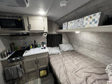 RV rental interior: compact kitchen (microwave, stove, sink), cozy bed with pillows, and overhead storage in neutral tones.