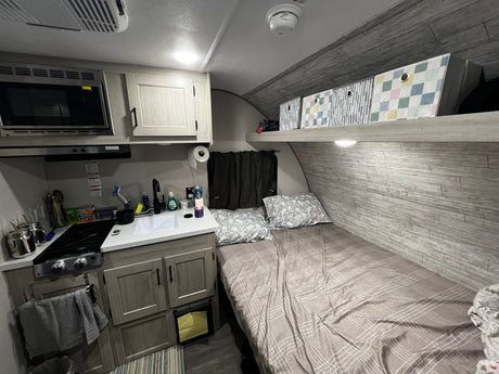 RV rental interior: compact kitchen (microwave, stove, sink), cozy bed with pillows, and overhead storage in neutral tones.