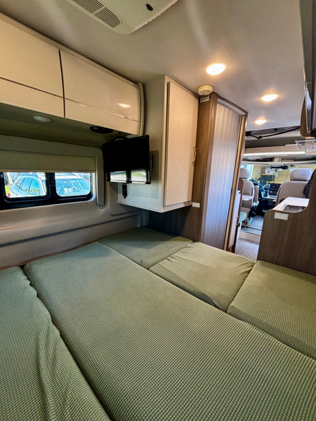 Cozy RV rental interior: green cushioned bed, windows, TV, overhead storage, and a view of the front cabin.