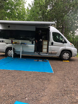 Person steps from a silver RV rental van at a gravel campsite. Outdoor table and blue tarp create a cozy setup.