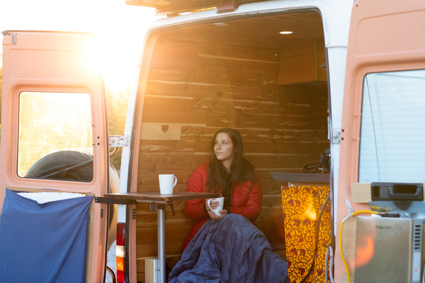 Woman bundled in blanket sips warm drink inside sunlit, wood-paneled RV rental van.