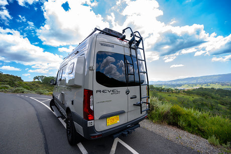 This Winnebago Revel Sprinter rv rental, parked on a scenic mountain road, offers stunning views of a green valley under a blue, cloudy sky.