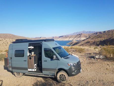 This grey RV rental is parked with its side door open, revealing its interior by a beautiful blue lake and arid mountains.