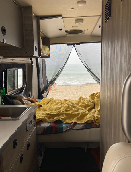 Inside an RV rental, a comfy bed and kitchenette face a stunning beach and ocean view, seen through a mesh screen.
