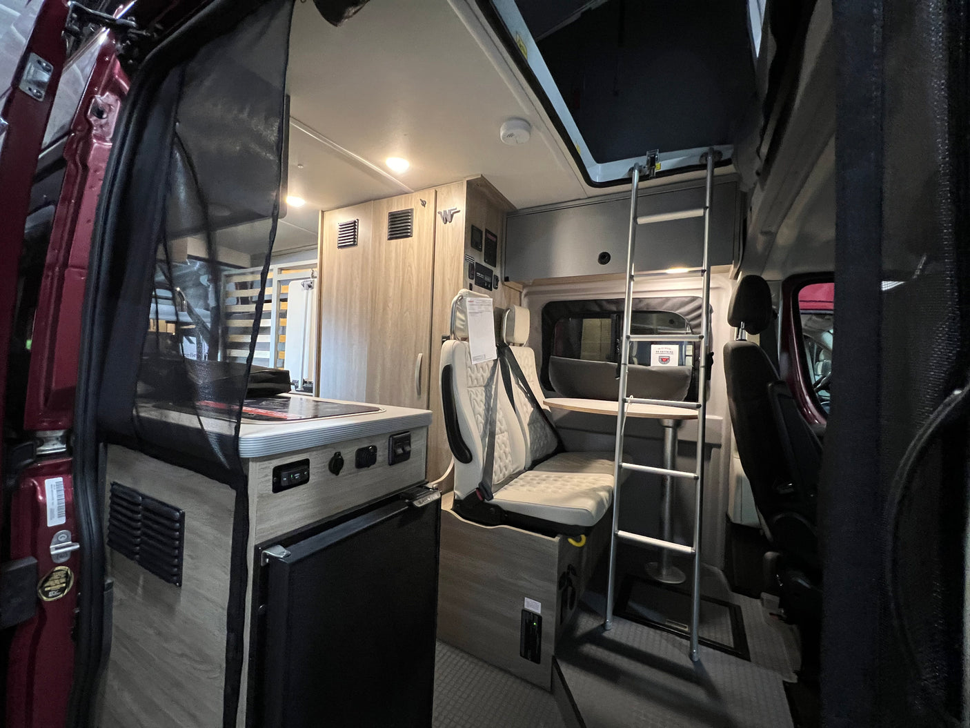 Interior of a modern RV rental, showcasing a compact kitchen, dinette, and ladder leading to a bunk bed for mobile living.