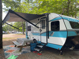 An RV rental with an extended awning, picnic table, and outdoor rug at a shaded campsite.