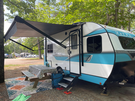 An RV rental with an extended awning, picnic table, and outdoor rug at a shaded campsite.