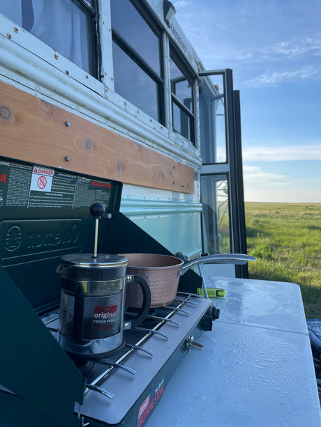 RV rental outdoor cooking: French press and pot on a Coleman propane stove next to a converted bus in a field.