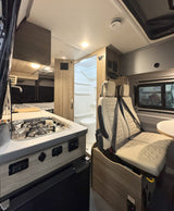 This RV rental showcases a compact camper van interior with a kitchenette, rear bed, bathroom, and front passenger seats.