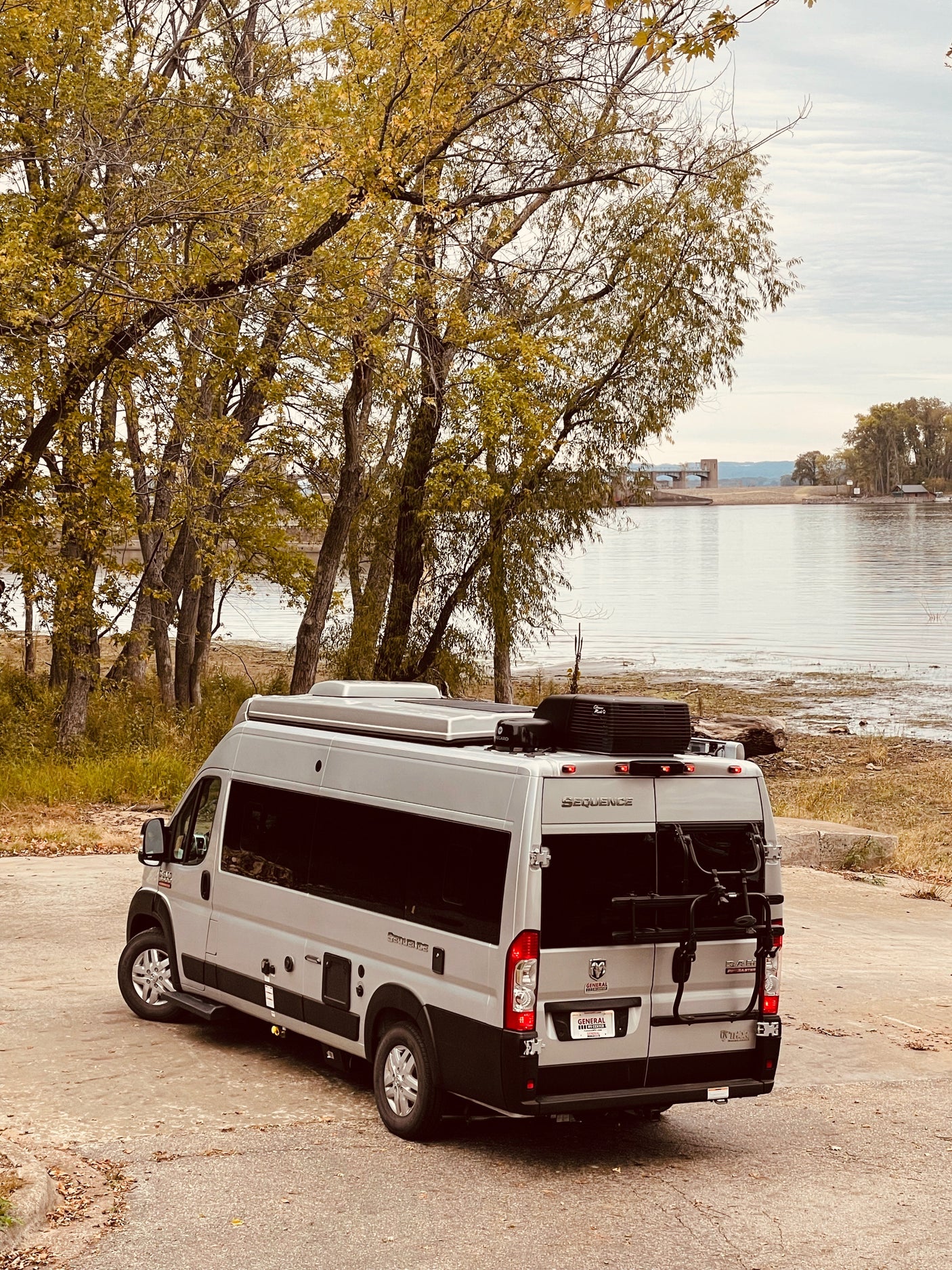 A modern silver RV rental parked on a paved bank, overlooking a serene river with autumn foliage.