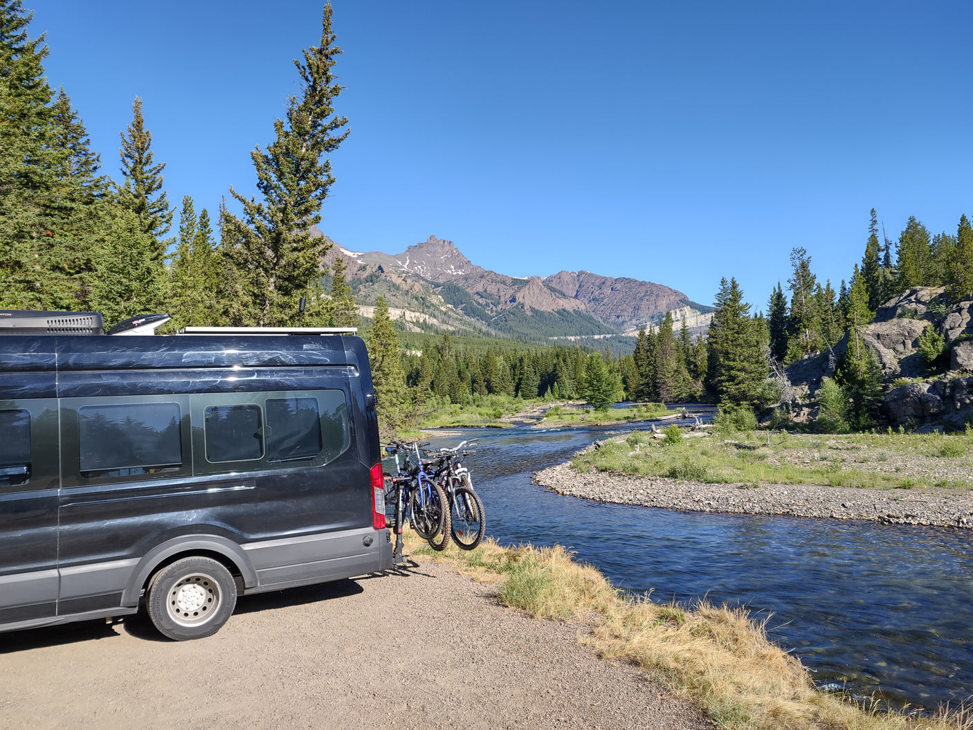 Dark RV rental camper van with bikes by a clear river, evergreen trees, and majestic mountains under a blue sky.
