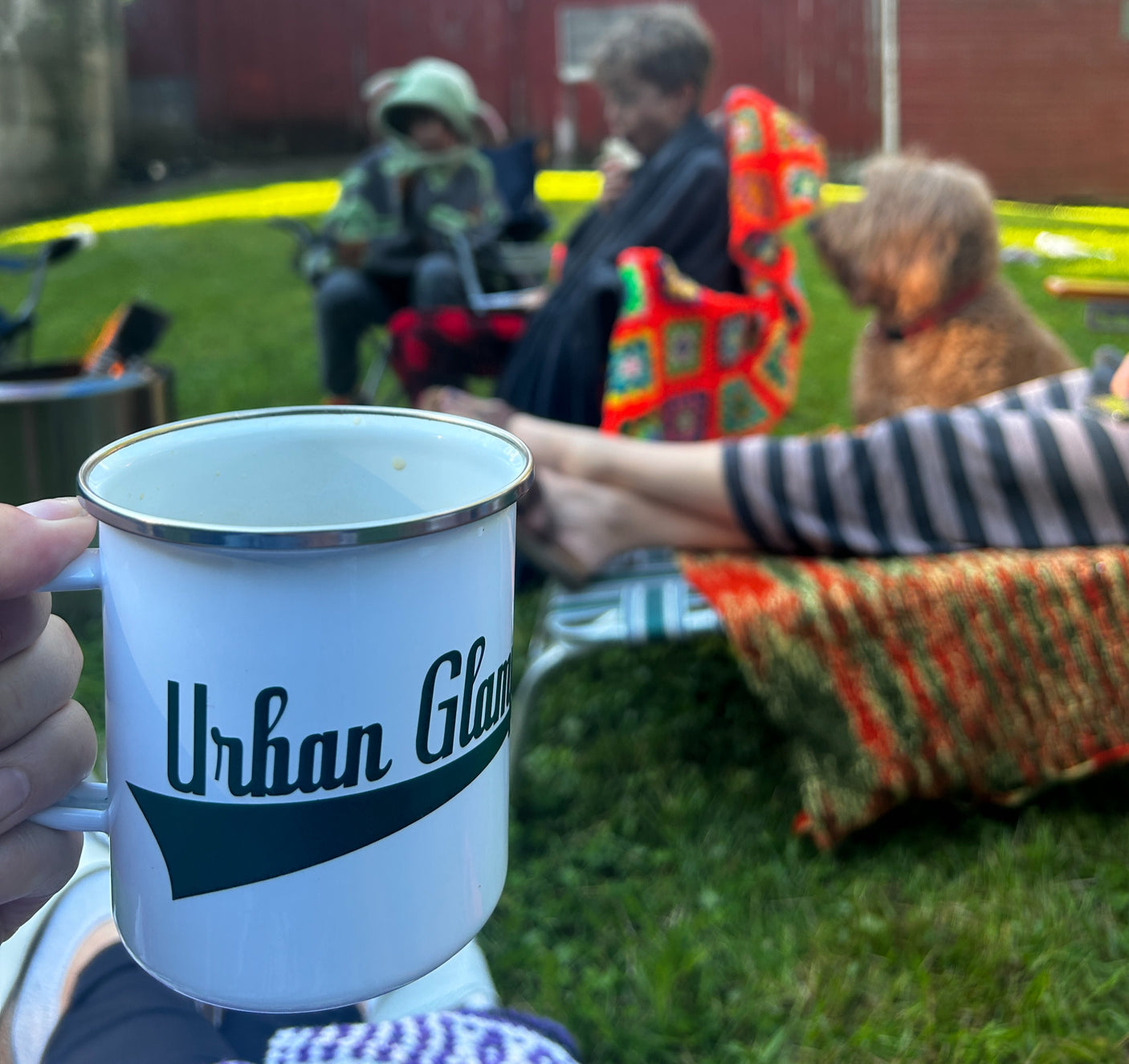 Outdoor relaxation with an 'Urban Glamp' mug, family, dog, and campfire. Great for RV rental adventures!