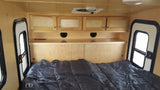 RV rental's light wood interior with bed, overhead storage, and side windows. A compact, cozy sleeping space.