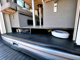 RV rental cargo space: neatly organized hose, box, and bag on a textured rubber floor under raised seating.