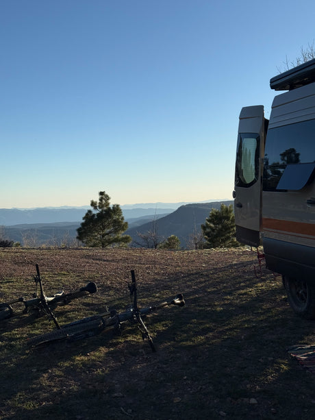 An RV rental is parked with its rear door open, overlooking mountains, with two mountain bikes on the ground under a clear blue sky.