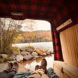 RV rental comfort: Plaid interior frames autumn's beauty. Feet in boots, a tranquil lake, & vibrant fall foliage.