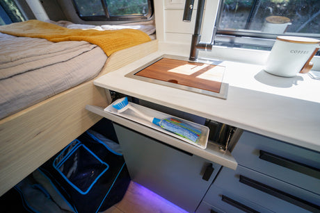 Inside a sleek RV rental: comfy bed, compact kitchen with a covered sink, and a clever pull-out tray for cleaning tools.
