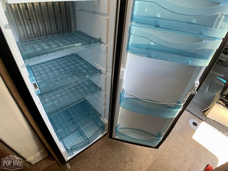 Inside an RV rental refrigerator, showing clean, empty interior with clear grid shelves & blue door bins, ready for groceries.