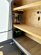 RV rental interior with wooden shelves, showcasing an inverter, battery charger, fuse box, and an E360 lithium battery.