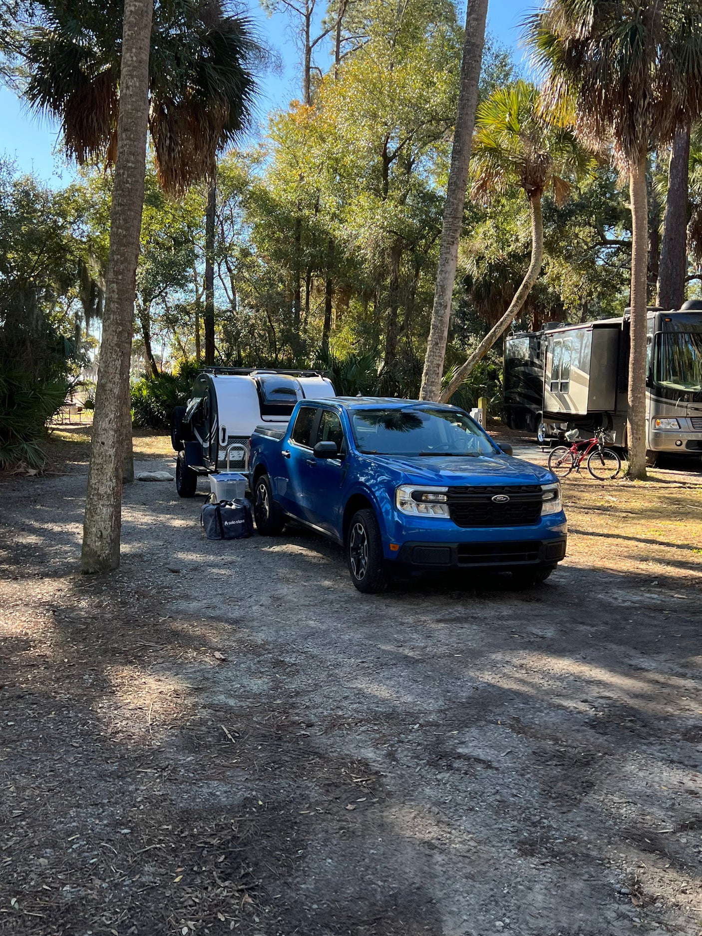 Blue truck with rv rental camper at a sunny campsite. Larger motorhomes and palm trees in this serene camping setting.