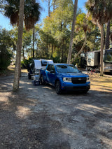 Blue truck with rv rental camper at a sunny campsite. Larger motorhomes and palm trees in this serene camping setting.